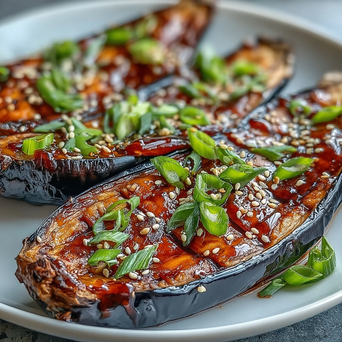 Miso Glazed Eggplant halves glistening with a sweet-savory caramelized glaze, garnished with sesame seeds and green onions on a white plate.