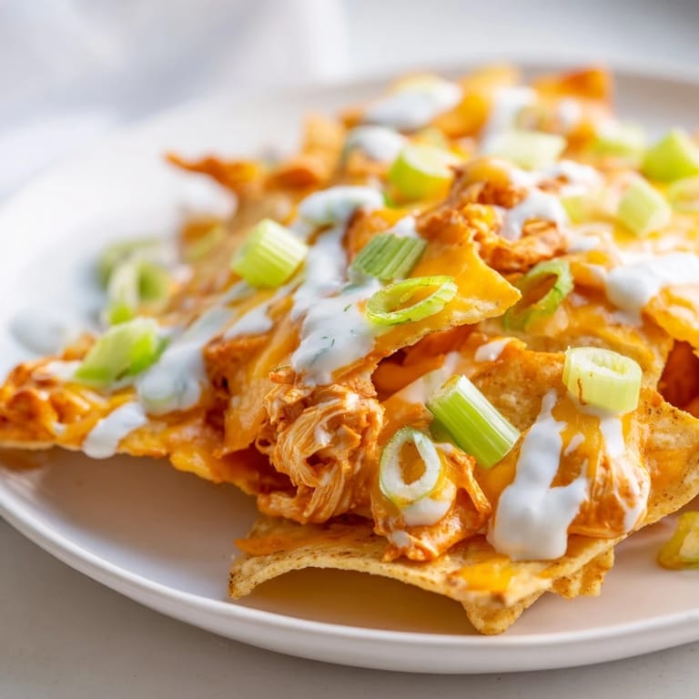 A vibrant serving of Buffalo Chicken Nachos on a platter, featuring golden, melted cheese bubbling over spicy chicken and chips, garnished with fresh green onions, celery, and tomatoes.
